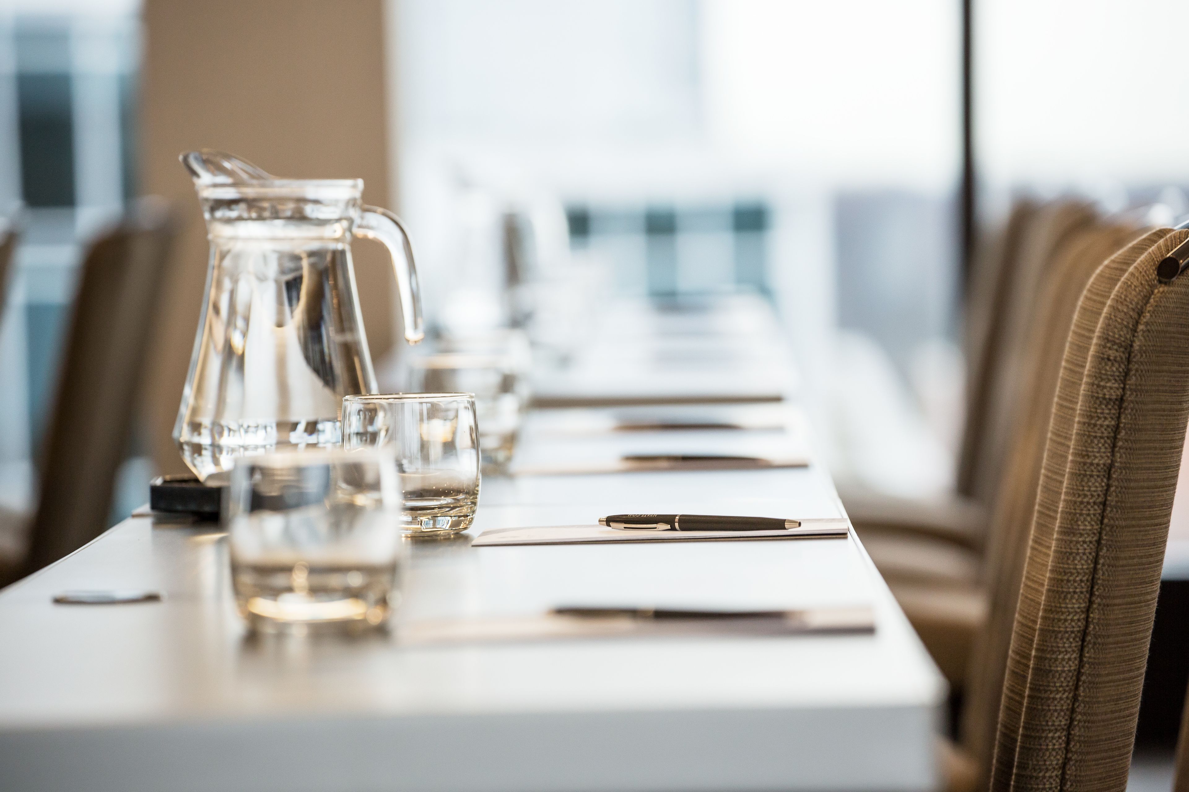 Una mesa de conferencias con una jarrón, vasos y bolígrafos, en un salón con ventanas amplias y vistas a edificios.