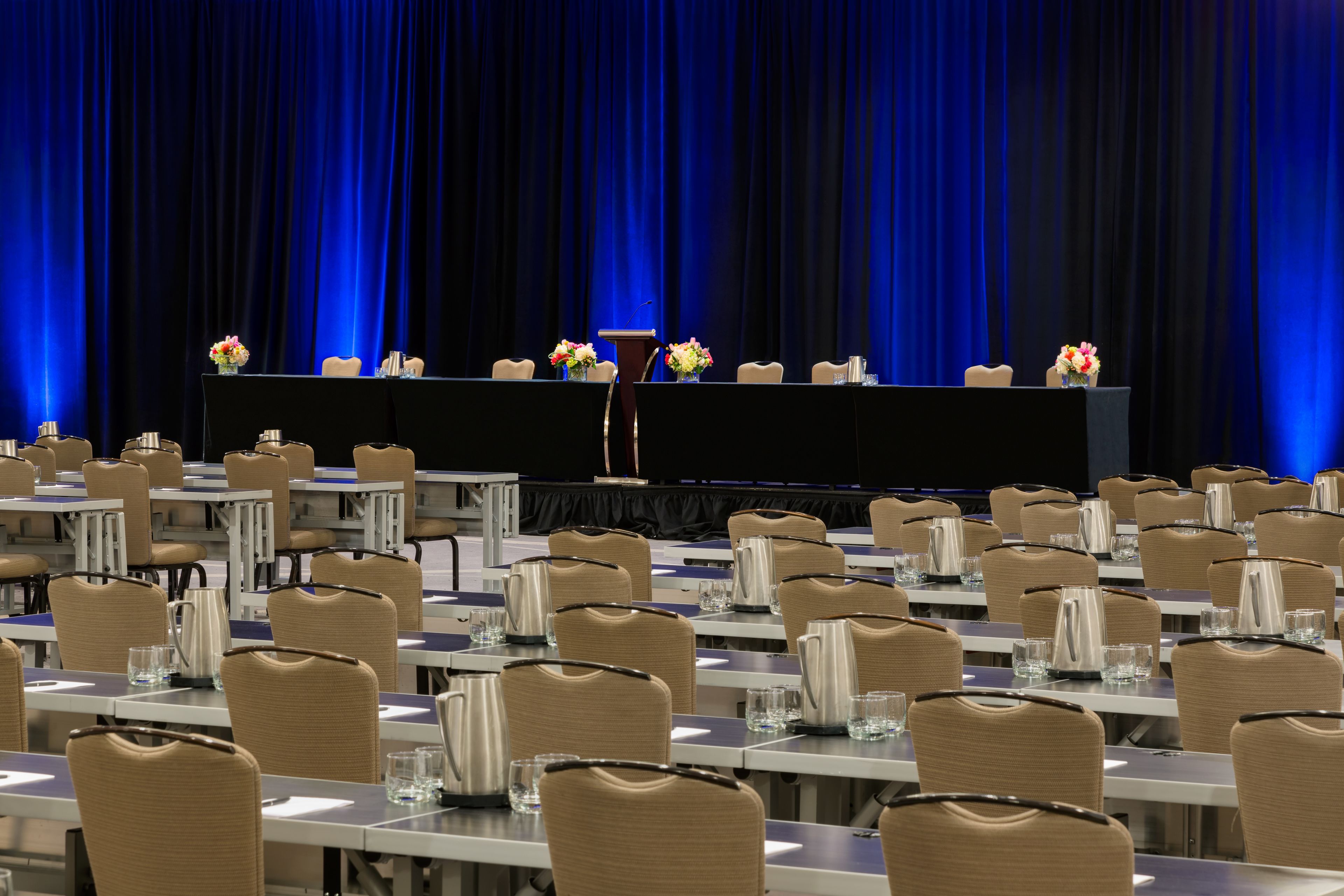 A conference room with rows of tables and chairs, a podium, and floral centerpieces.
