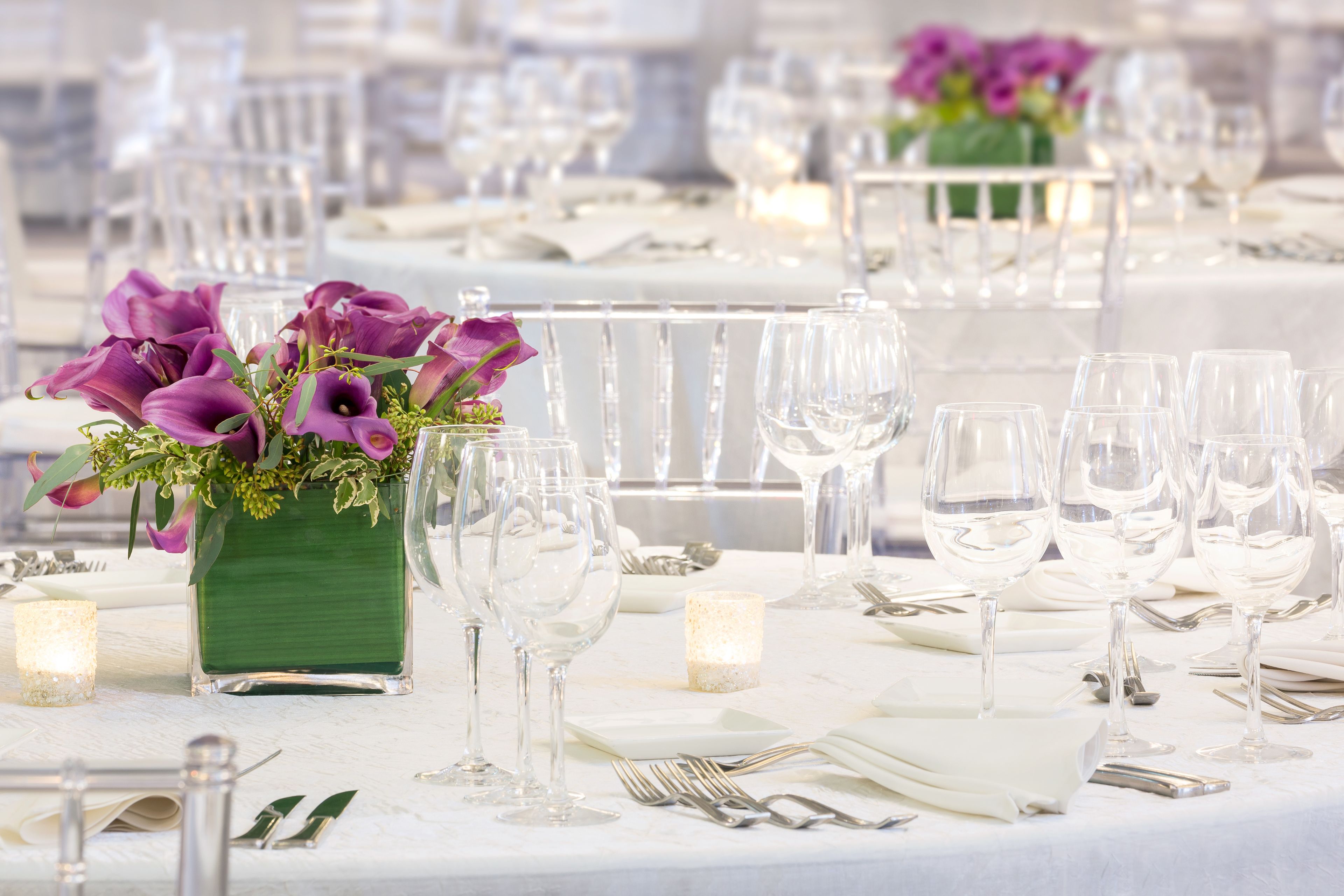 A banquet table set with white tablecloths, clear glassware, and a centerpiece of purple flowers in a green vase.