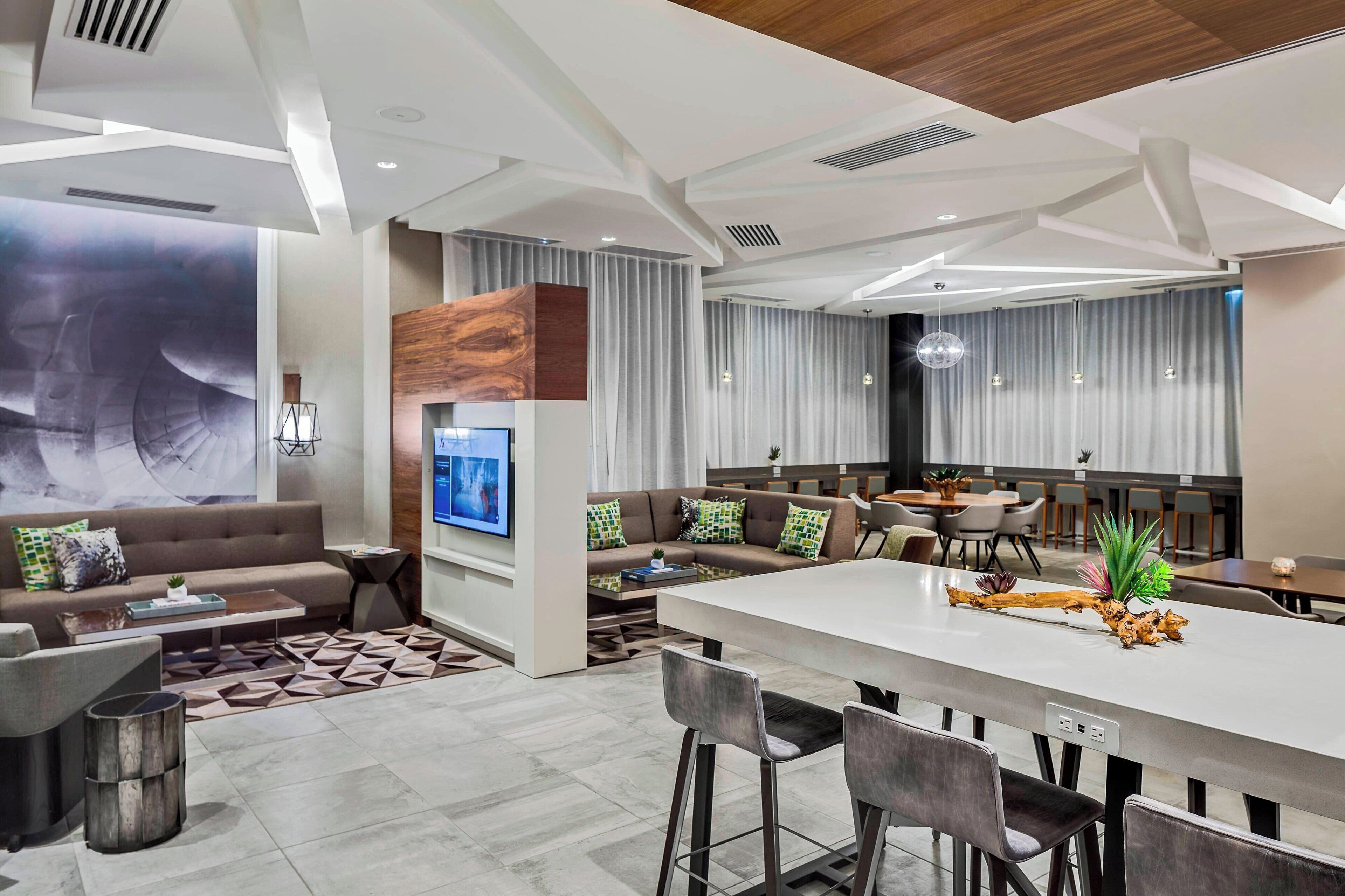 A modern hotel lobby with a central bar area, seating arrangements, and a large wall-mounted TV.