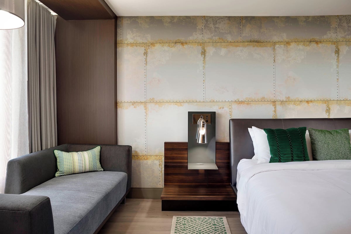 A modern hotel room with a sofa, a bed, a nightstand, and a wall-mounted light fixture.