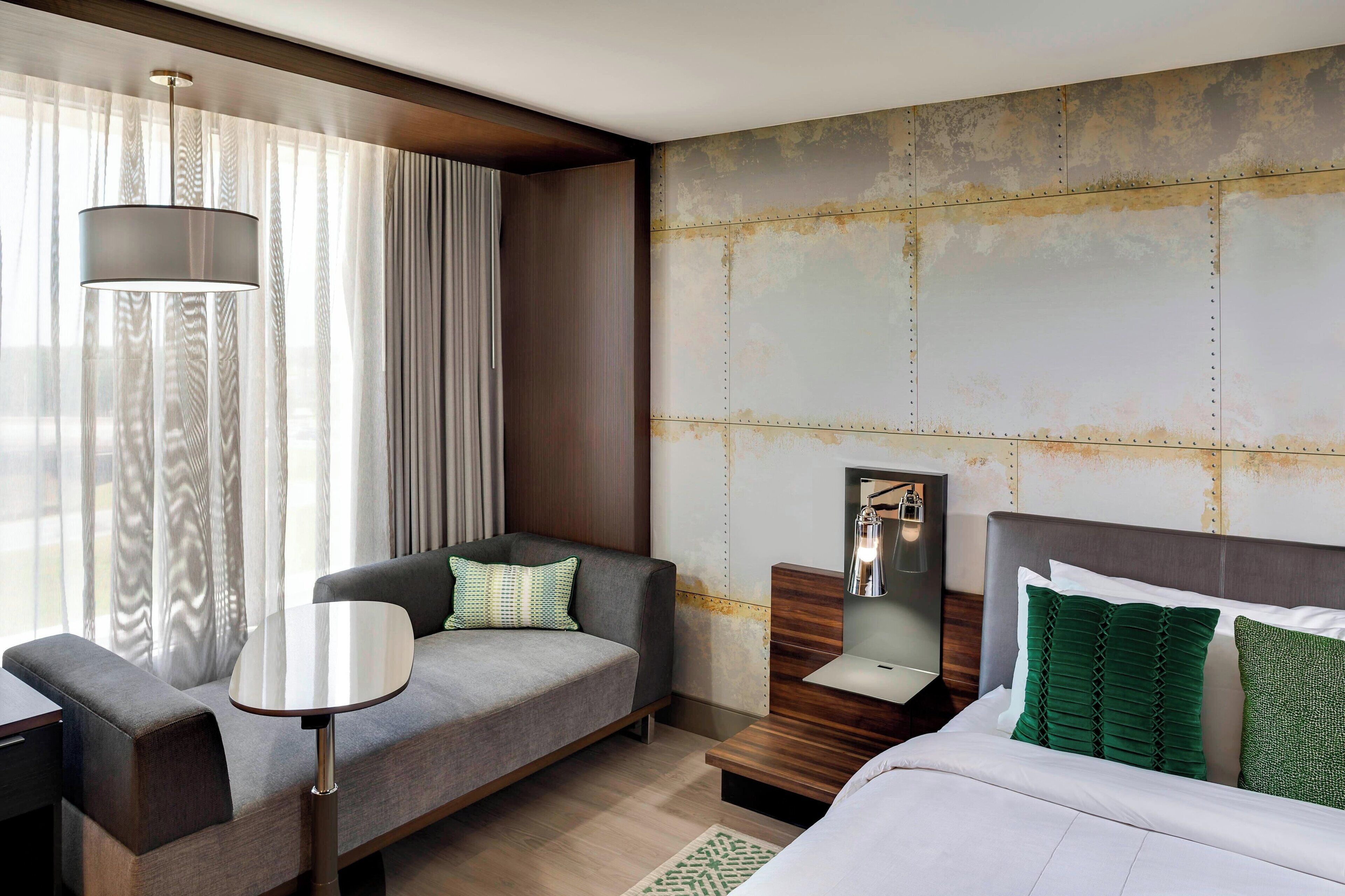 Une chambre d’hôtel moderne, équipée d’un canapé, d’une petite table, d’une table de chevet avec une lampe et d’un lit avec des oreillers verts.