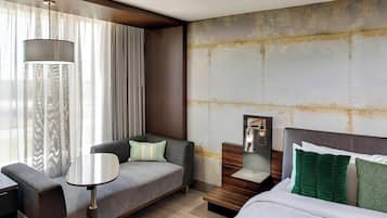 Une chambre d’hôtel moderne, équipée d’un canapé, d’une petite table, d’une table de chevet avec une lampe et d’un lit avec des oreillers verts.