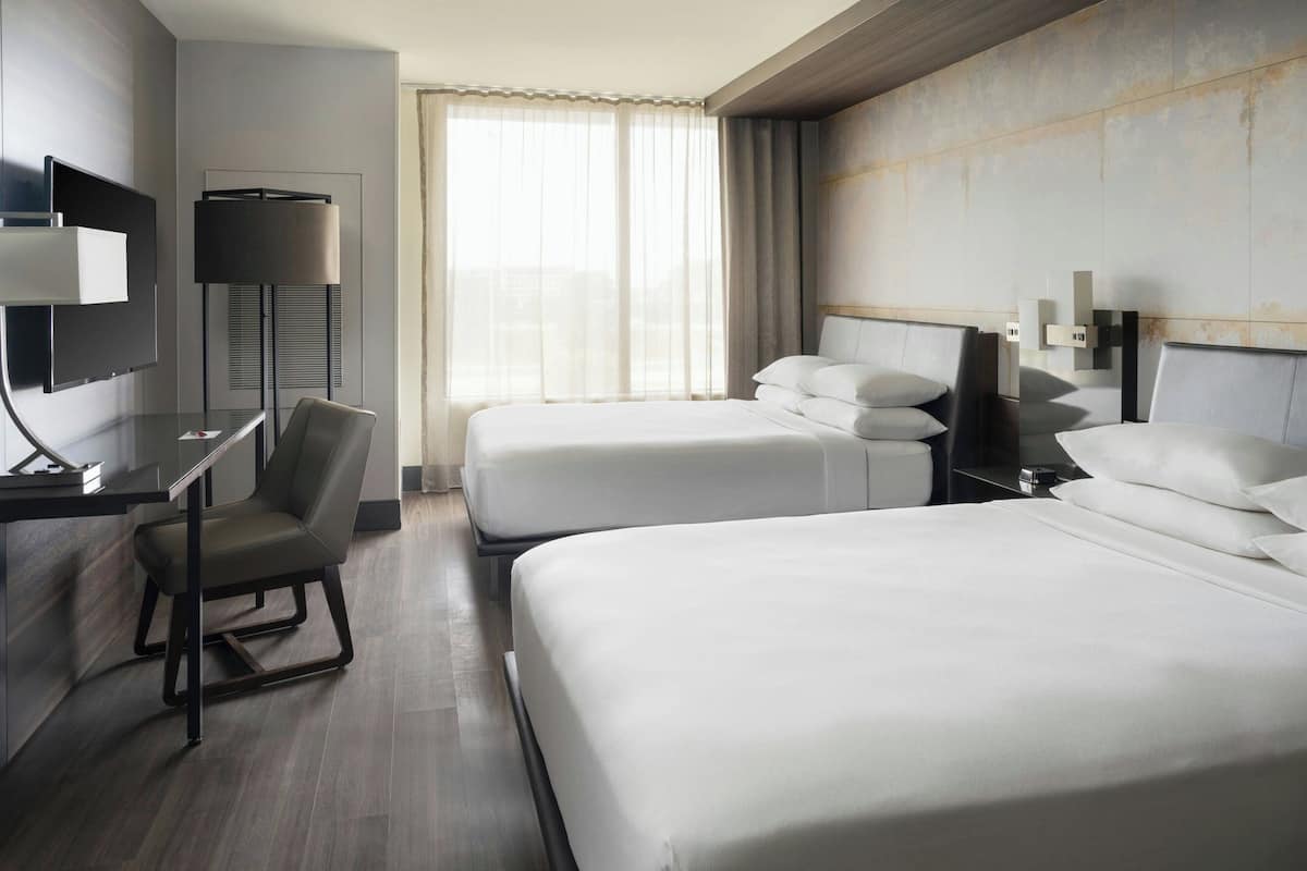 A hotel room with two beds, a desk, a chair, a TV, and a large window with curtains.