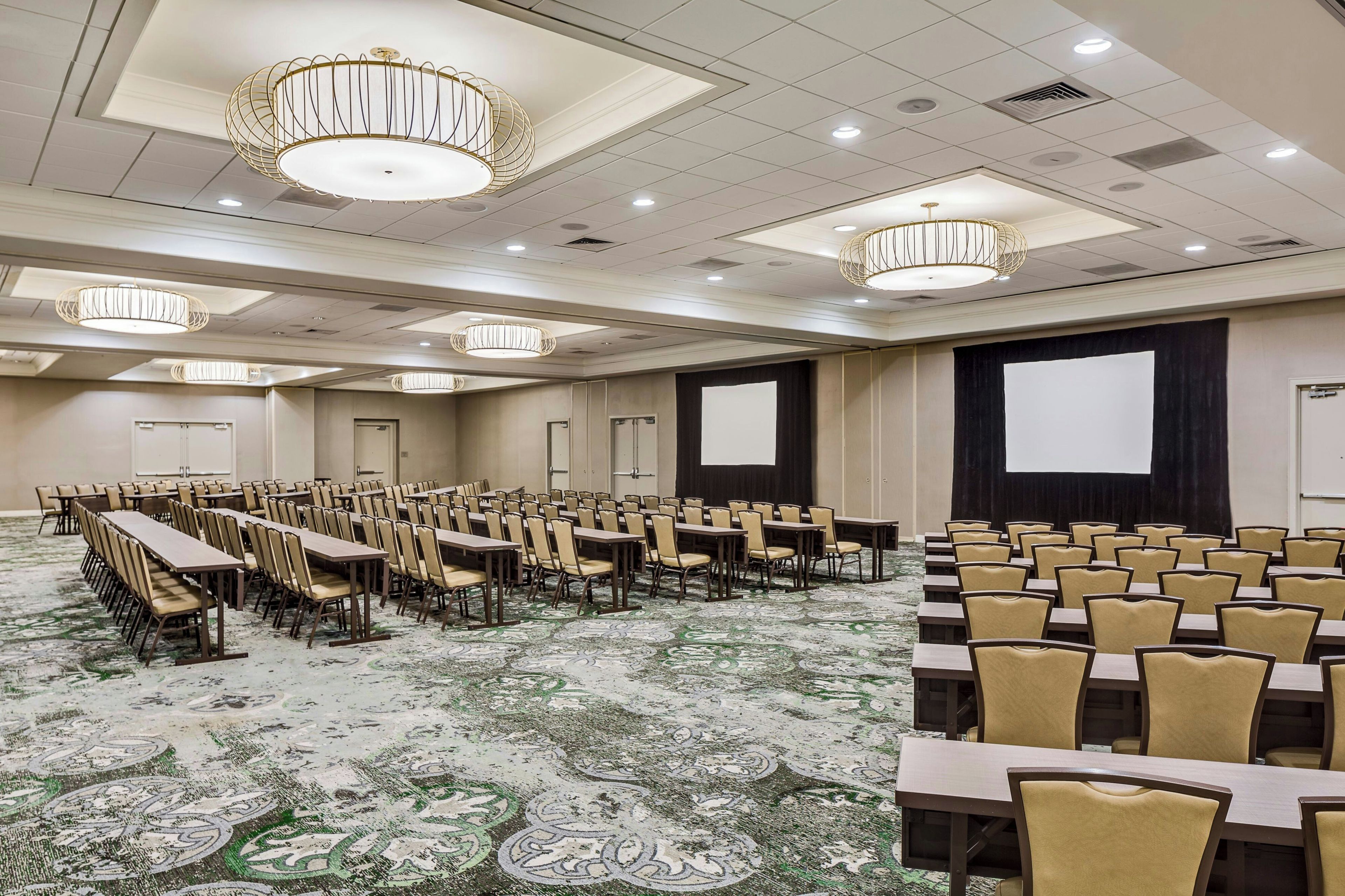 Un salón de conferencias con filas de mesas y sillas, grandes candelabros y una pantalla para proyector.
