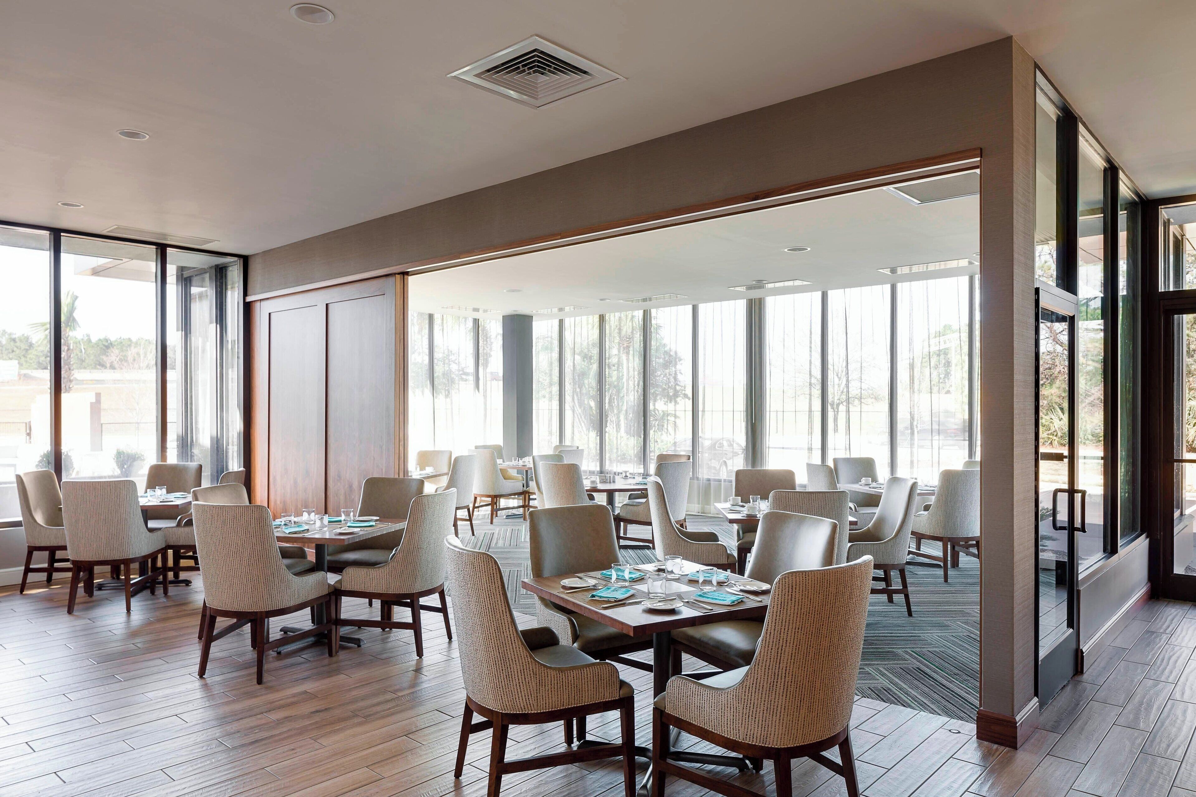 Un amplio comedor con múltiples mesas y sillas, ventanas grandes que aportan luz natural, y un diseño de interiores moderno.