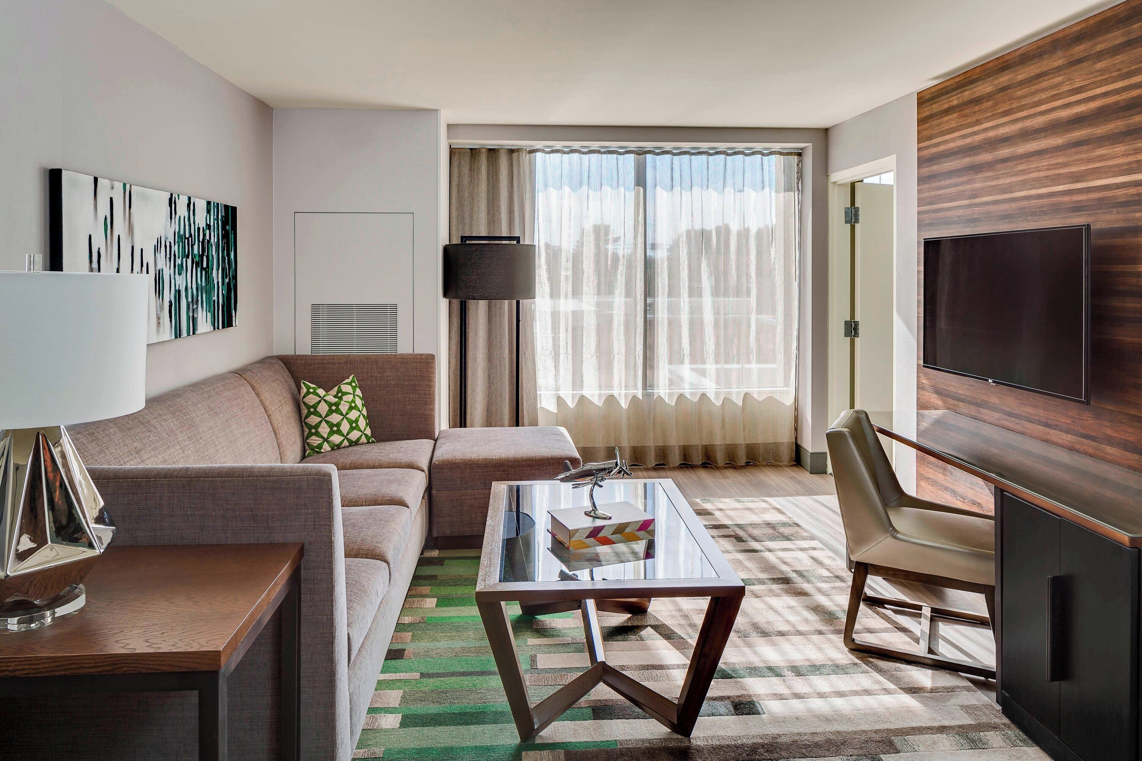 Une chambre d’hôtel moderne, dotée d’un canapé, d’une table basse en verre, d’un téléviseur et d’une grande fenêtre avec des rideaux.