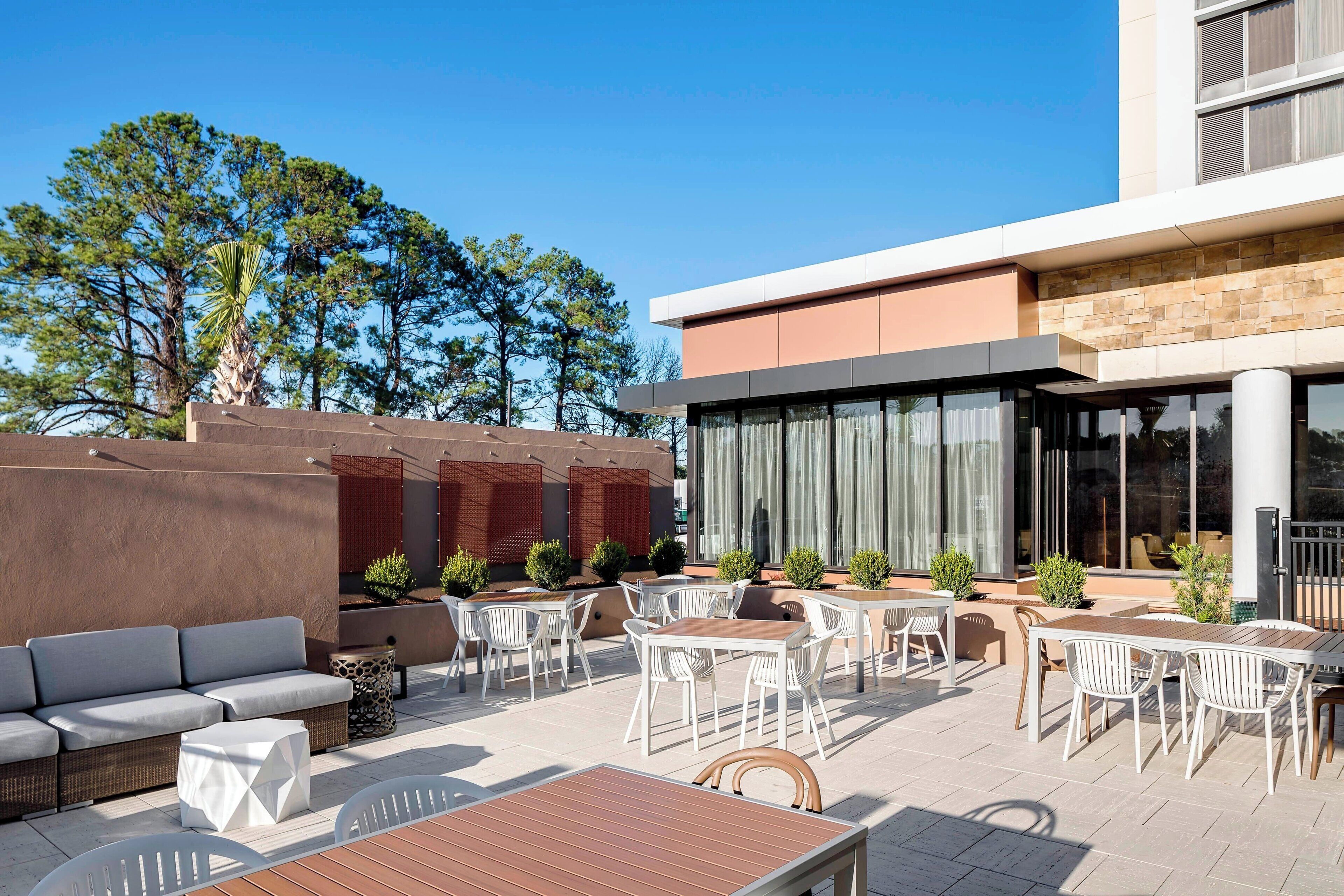 An outdoor patio with seating and tables, surrounded by a modern building with large windows and a stone facade.
