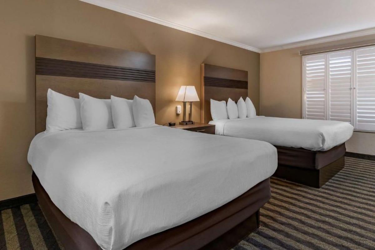 Une chambre d’hôtel avec deux lits, une table de chevet, une lampe et une fenêtre avec des stores.