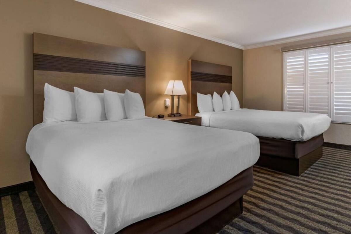 A hotel room with two beds, a nightstand, a lamp, and a window with blinds.