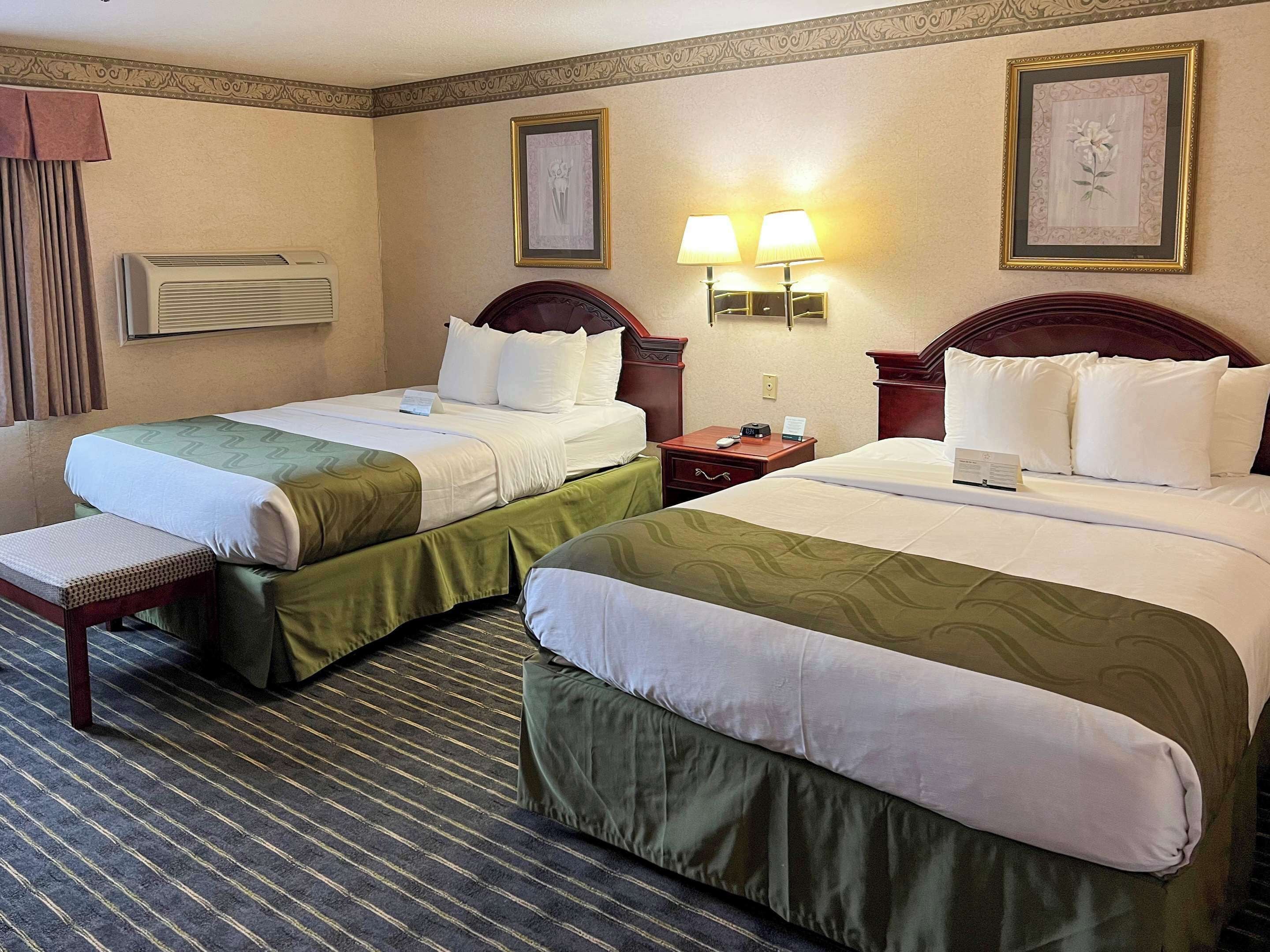 A hotel room with two beds, each with a green and white bedspread, a bedside table with a lamp, and a wall-mounted air conditioner.