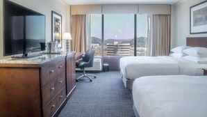 Habitación de hotel con un televisor grande, un escritorio con lámpara, dos camas y vistas a un paisaje urbano a través de amplias ventanas.
