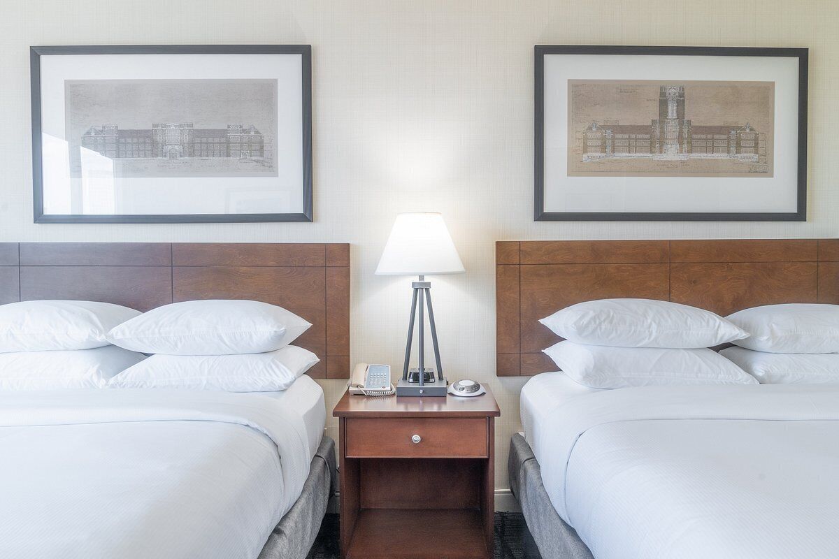 Ein Hotelzimmer mit zwei Betten, einem Nachttisch mit Lampe und gerahmten architektonischen Zeichnungen an der Wand.