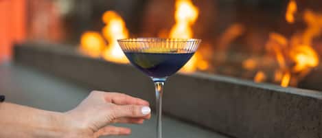 Una mano sostiene una copa de martini con una bebida azul oscuro, sobre un fondo borroso de fuego.