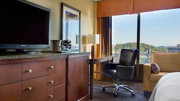 A hotel room with a flat-screen TV, a desk with a lamp, a chair, a bed, and a large window with a city view.