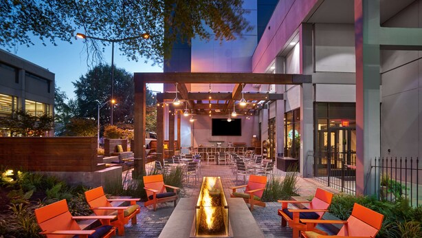 Un patio al aire libre con sillas naranjas, una chimenea central y una pérgola con luces colgantes.