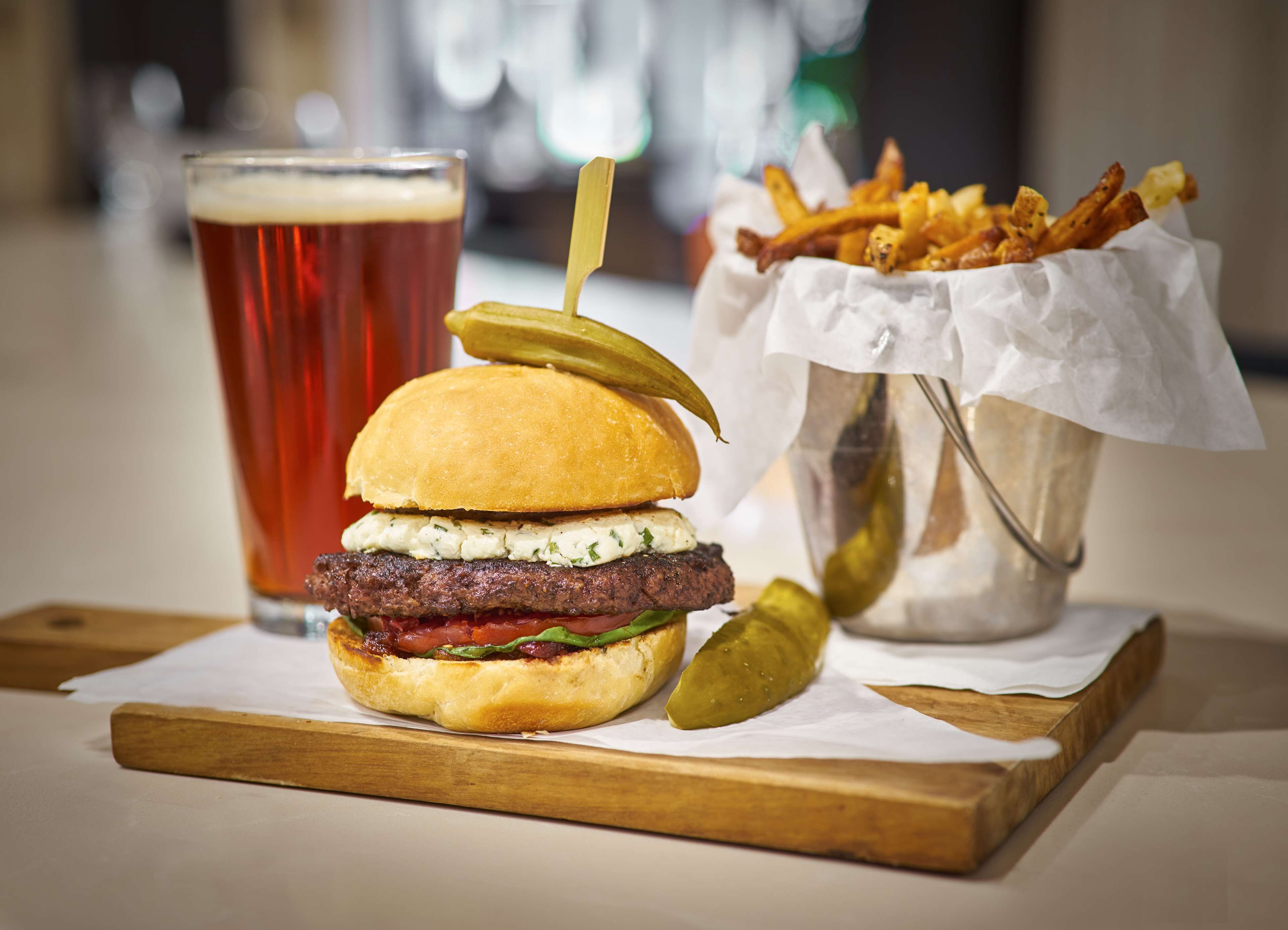 Un hamburger con un sottaceto, un bicchiere di birra e un cestino di patatine fritte su un tagliere di legno.