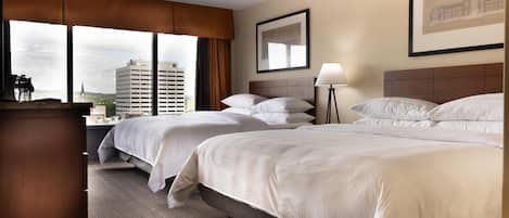 Habitación de hotel con dos camas, un ventanal con vistas a un paisaje urbano, un cuadro en la pared y una lámpara en la mesita de noche.