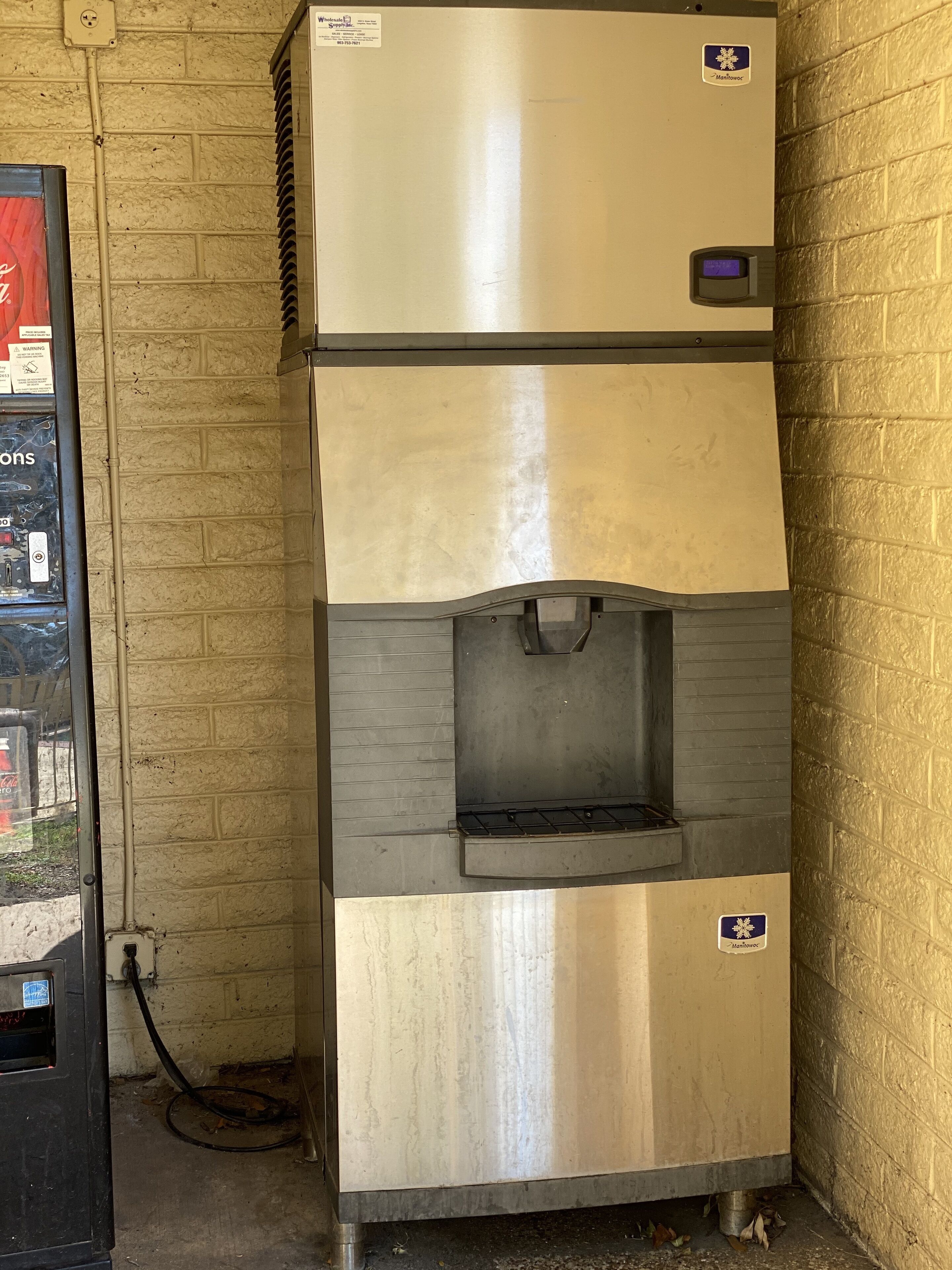 A stainless steel commercial refrigerator with a water dispenser, located in a corner against a brick wall.