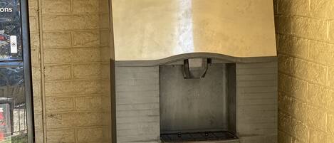 A stainless steel commercial refrigerator with a water dispenser, located in a corner against a brick wall.