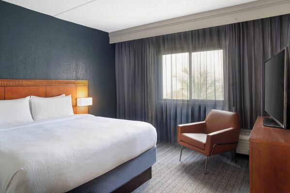 Una habitación de hotel con una cama grande, una silla, un televisor y un ventanal con cortinas.
