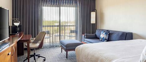 A hotel room with a bed, a desk with a chair, a television, and a large window with curtains.