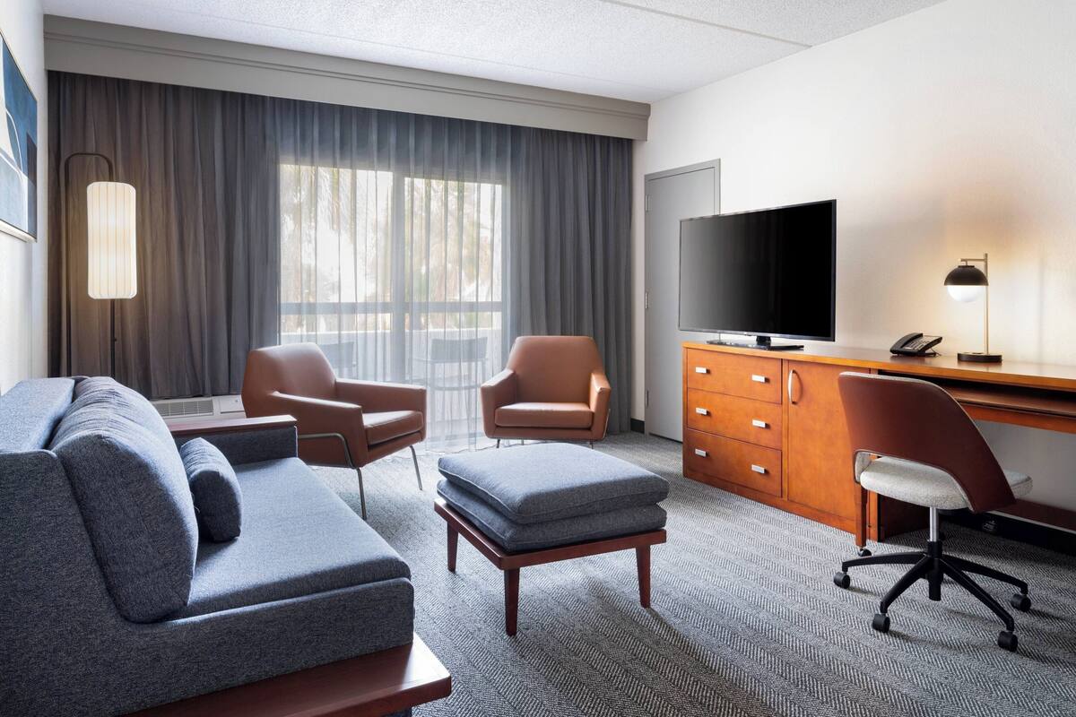 Ein Hotelzimmer mit einer Couch, zwei Sesseln, einem Schreibtisch mit Fernseher, einer Lampe und einem großen Fenster mit Vorhängen.