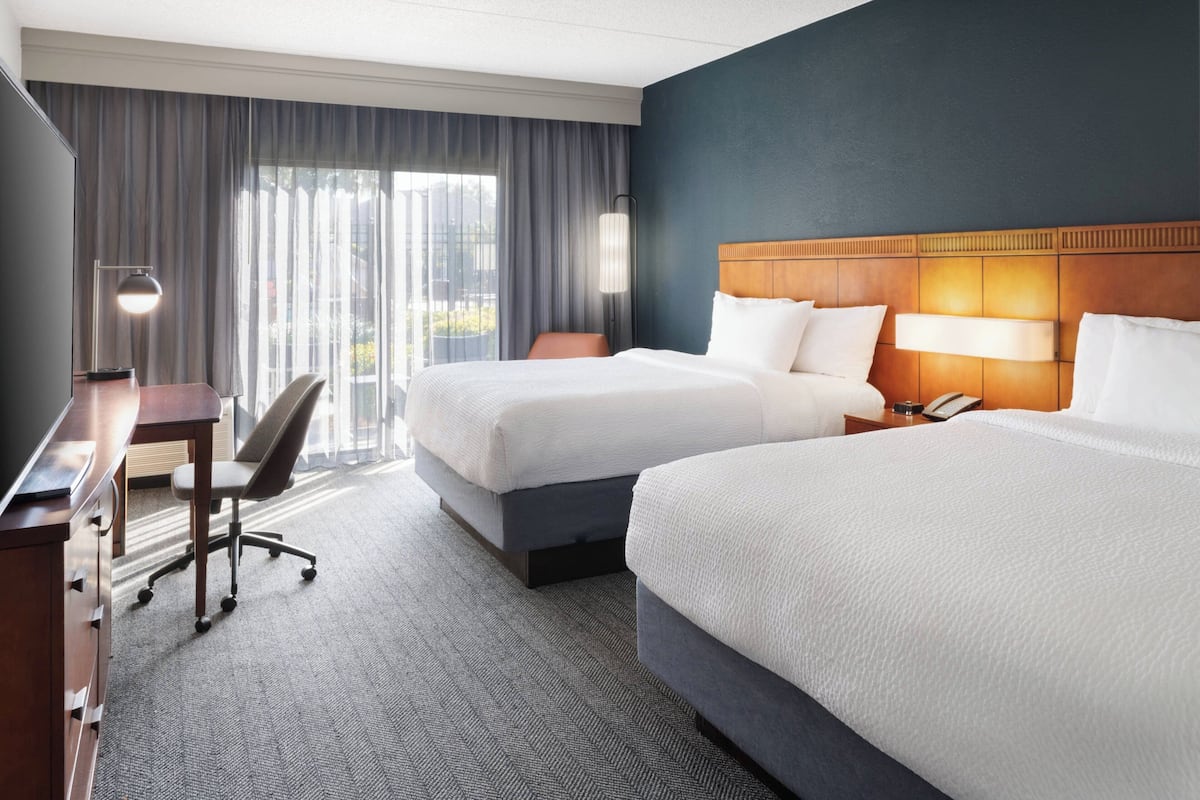 Ein Hotelzimmer mit zwei Betten, einem Schreibtisch, einem Stuhl und einem großen Fenster mit Vorhängen.