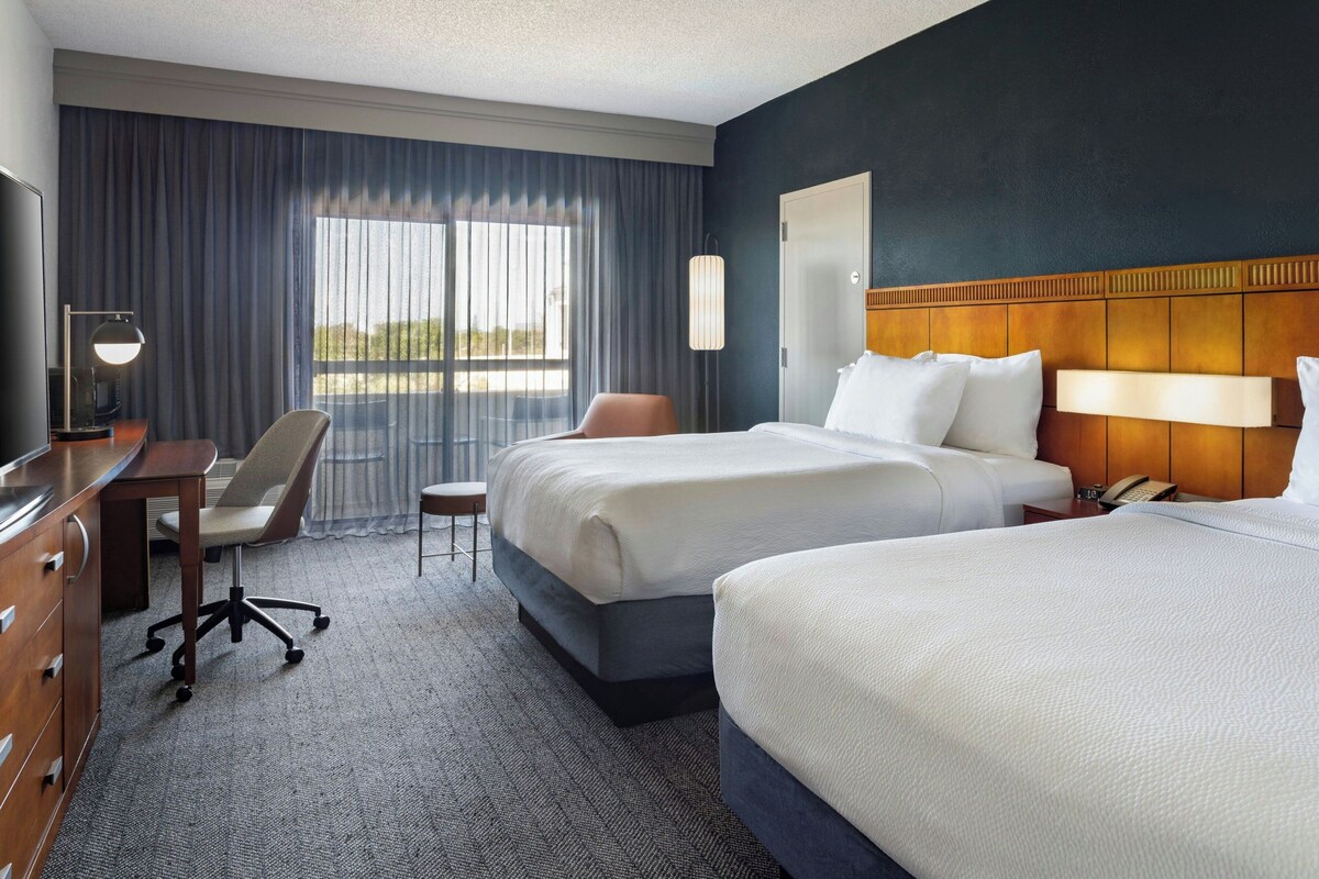A hotel room with two beds, a desk, a chair, a lamp, and a view of the outdoors.