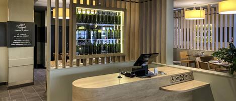Una recepción con mostrador de madera, exhibición de vinos detrás de un panel de madera, y un área de estar con mesas y sillas.