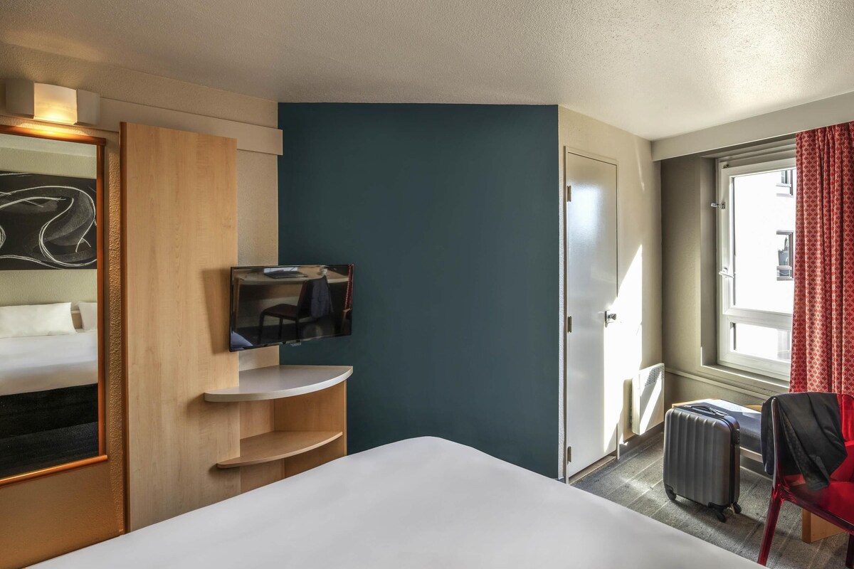 A hotel room with a bed, a TV mounted on a wooden shelf, a desk, a chair, a suitcase, and a window with curtains.