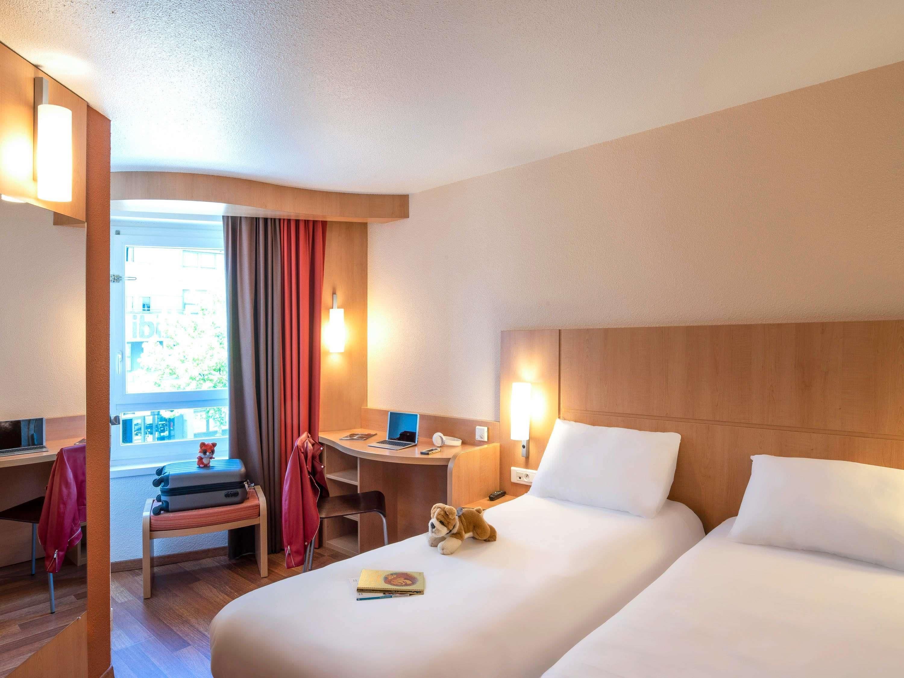 A hotel room with two beds, a desk, a chair, a laptop, and a window with curtains.
