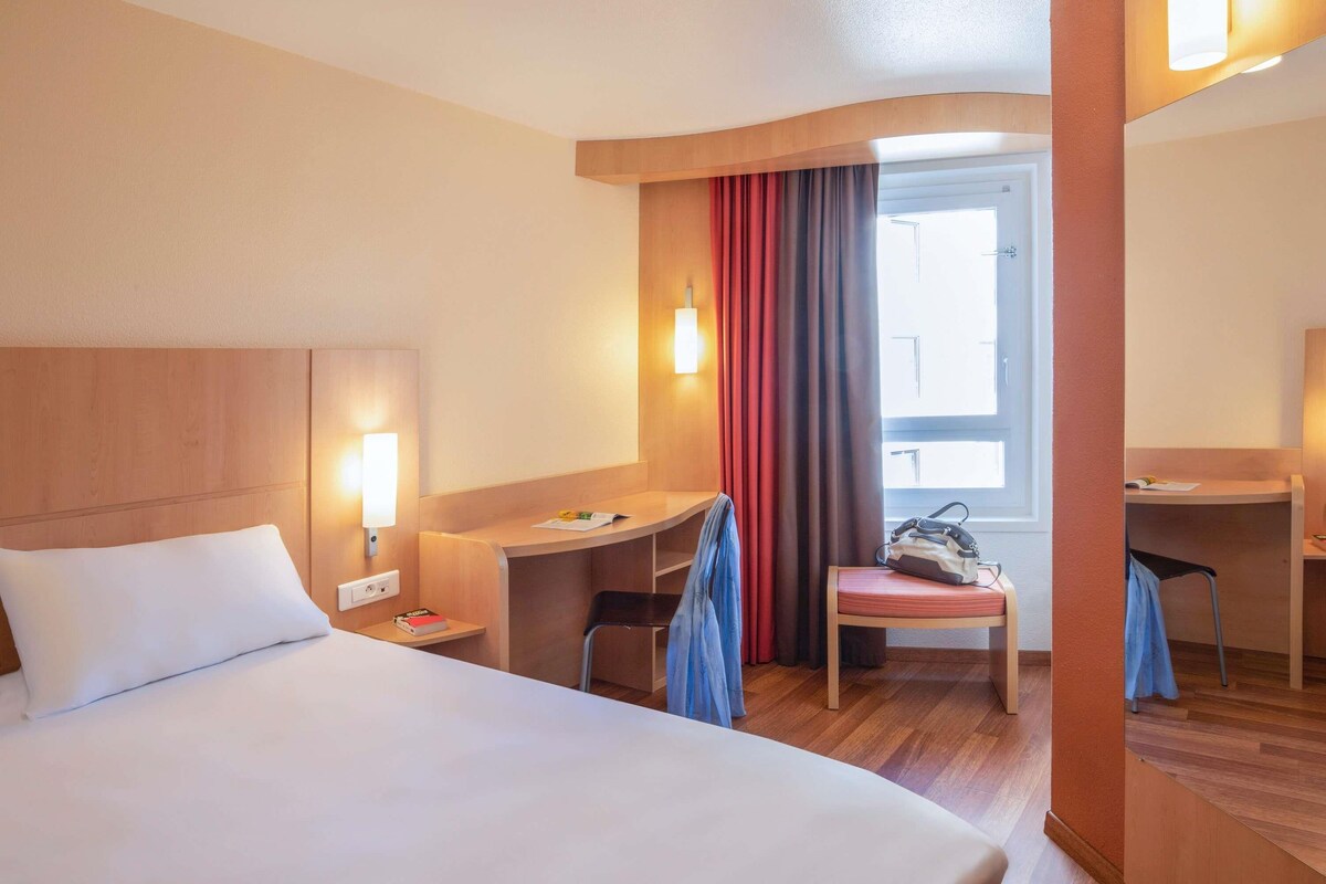 A hotel room with a bed, a desk, a chair, and a window with curtains.