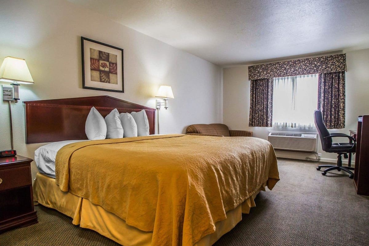 A hotel room with a large bed, two bedside lamps, a desk with a chair, an air conditioning unit, and a window with curtains.