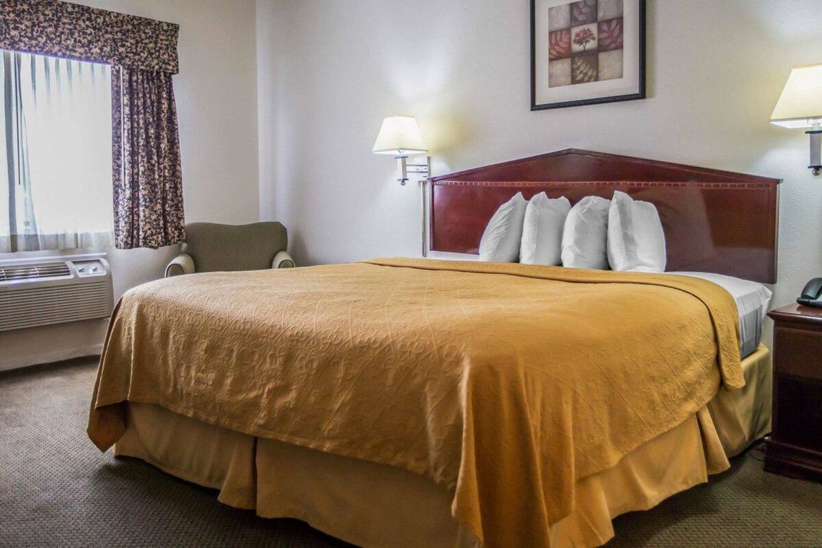 A hotel room with a large bed, two bedside lamps, a headboard, a framed picture on the wall, a window with curtains, and an air conditioning unit.