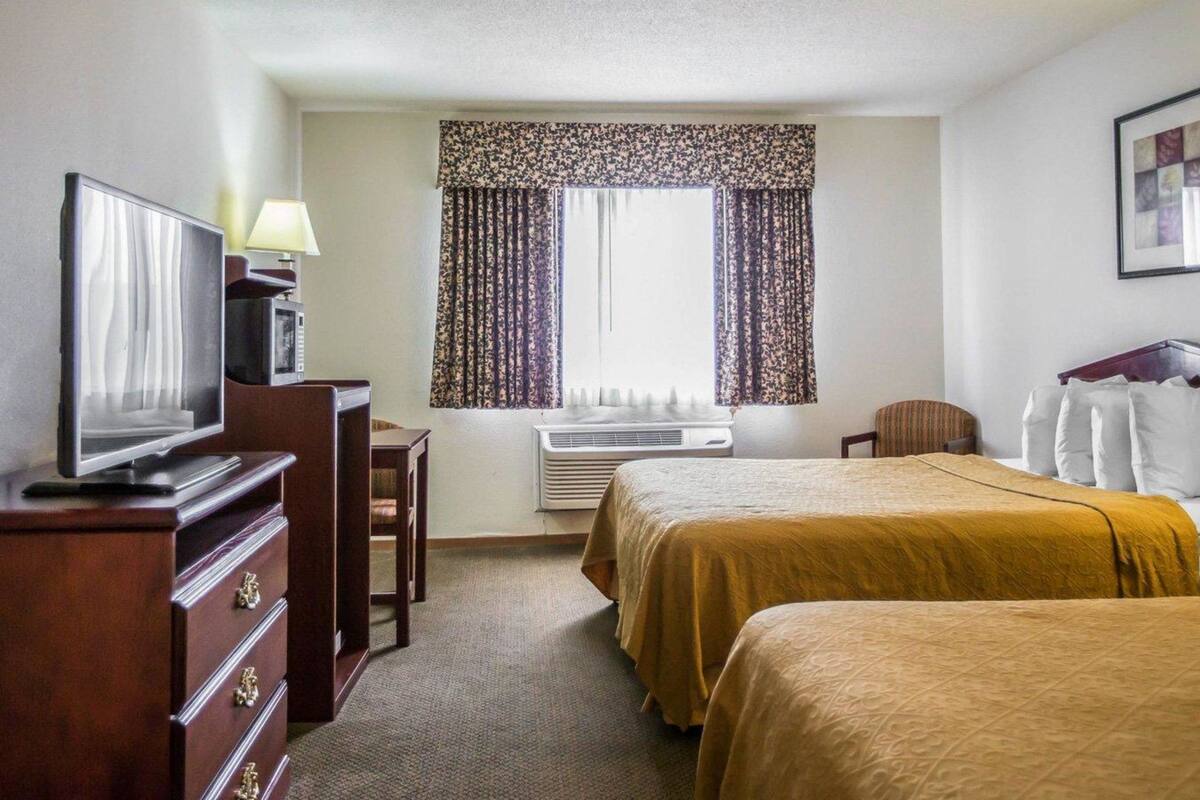 A hotel room with two beds, a desk with a television, a chair, and a window with curtains.