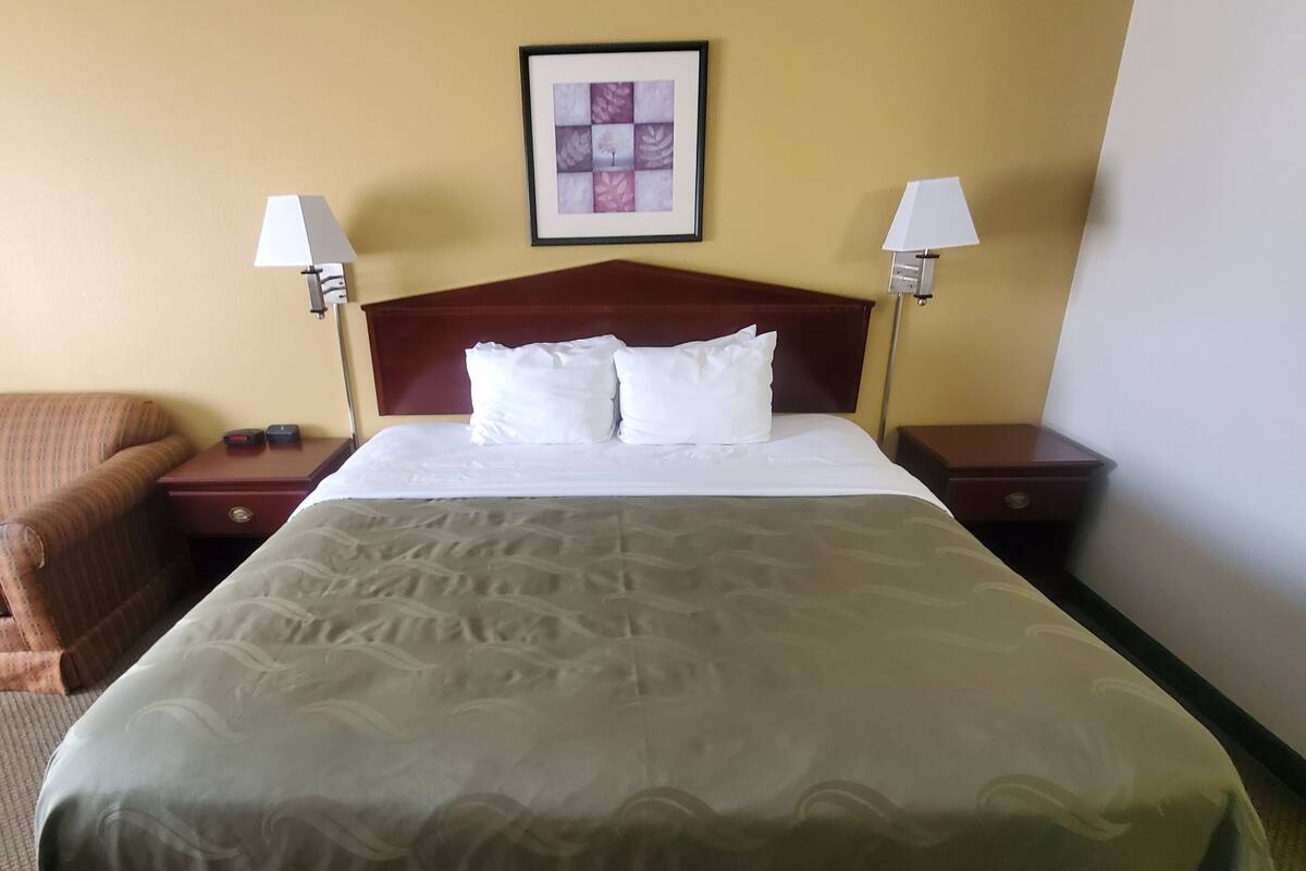 A hotel room with a large bed, two bedside tables with lamps, a framed picture on the wall, and a brown armchair.