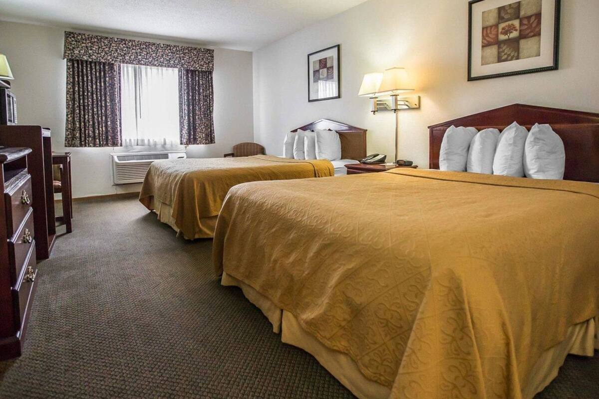 A hotel room with two beds, a desk, a chair, a lamp, and a window with curtains.