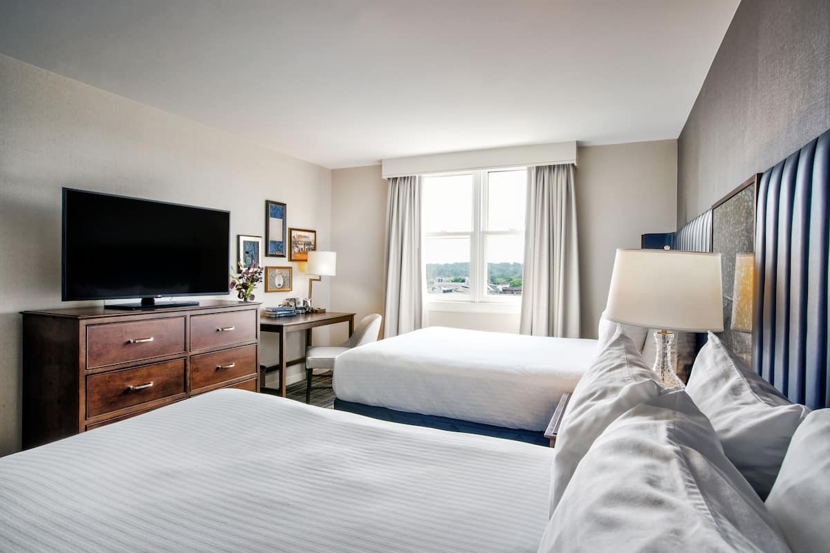 A hotel room with two beds, a desk, a television, and a window with curtains.