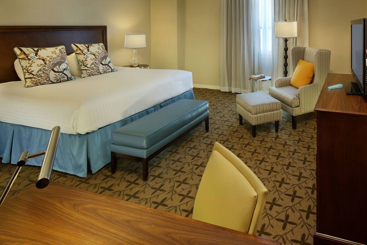 A hotel room with a bed, a desk, a chair, a bench, and a television.