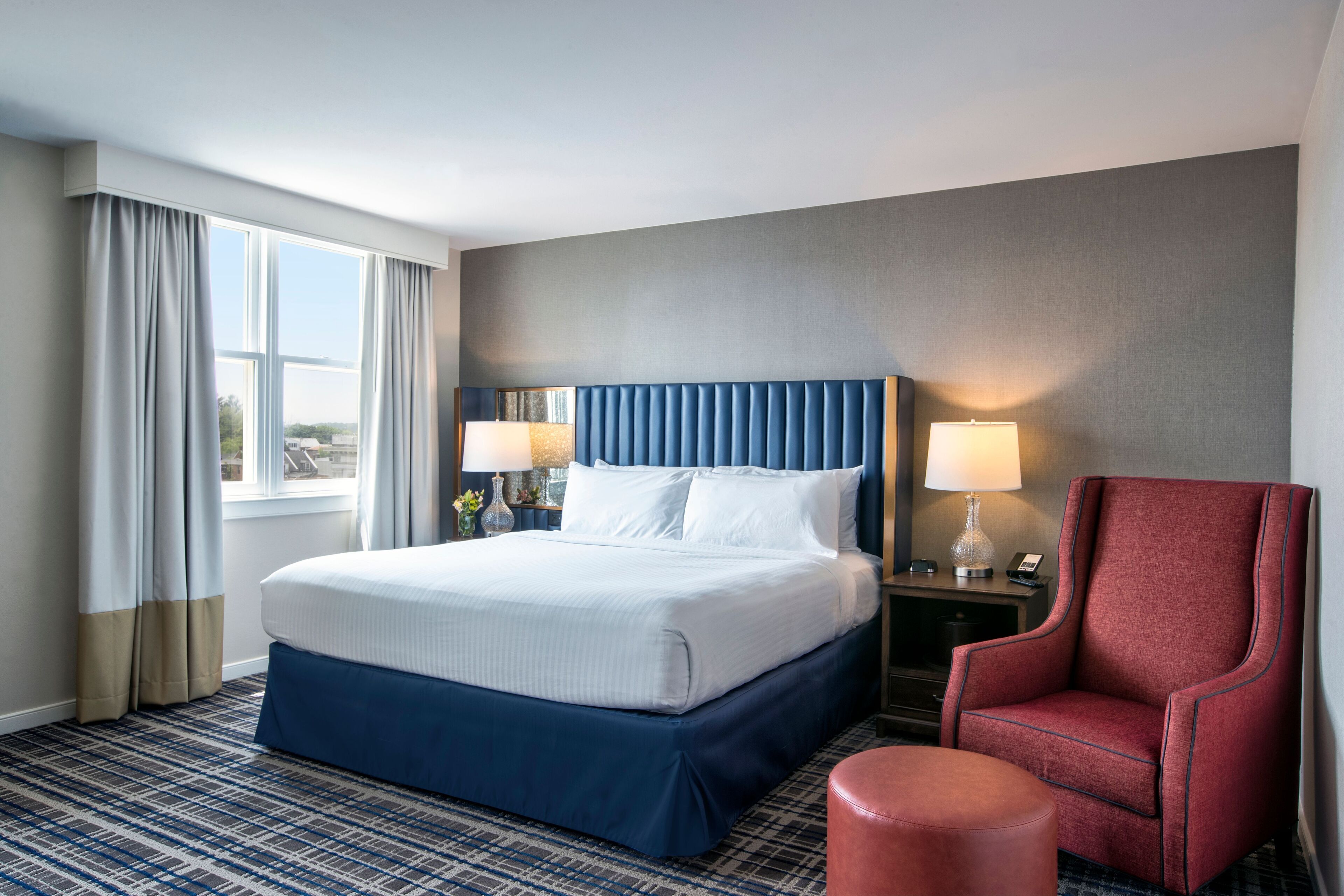 A hotel room with a large bed, two bedside tables with lamps, a red armchair, and a view of the outdoors.