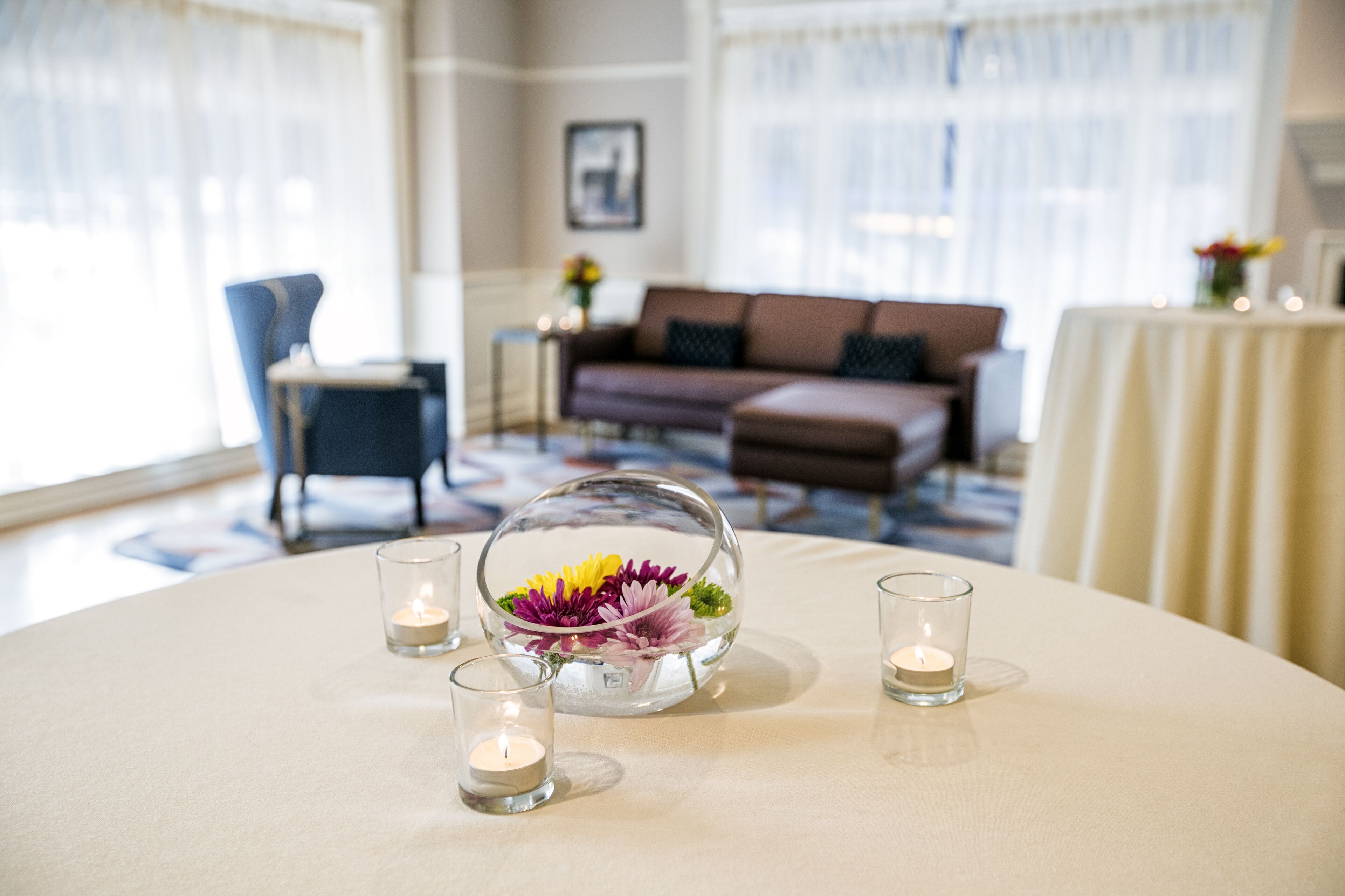A well-lit room with a round table, a glass dome centerpiece with flowers, and candles.