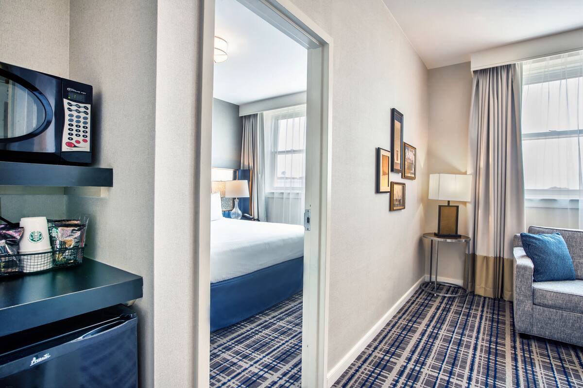 A hotel room with a bed, a desk with a microwave, a coffee maker, and a trash bin. There is a window with curtains and a view of the outside.