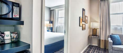 A hotel room with a bed, a desk with a microwave, a coffee maker, and a trash bin. There is a window with curtains and a view of the outside.
