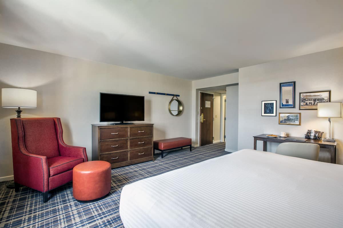 A hotel room with a large bed, a red armchair, a wooden dresser, a television, and a desk with a lamp.