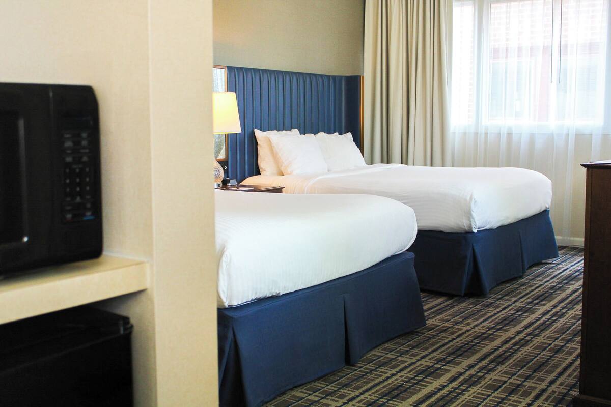 A hotel room with two beds, a desk with a lamp, a microwave, and a view of the city through the window.