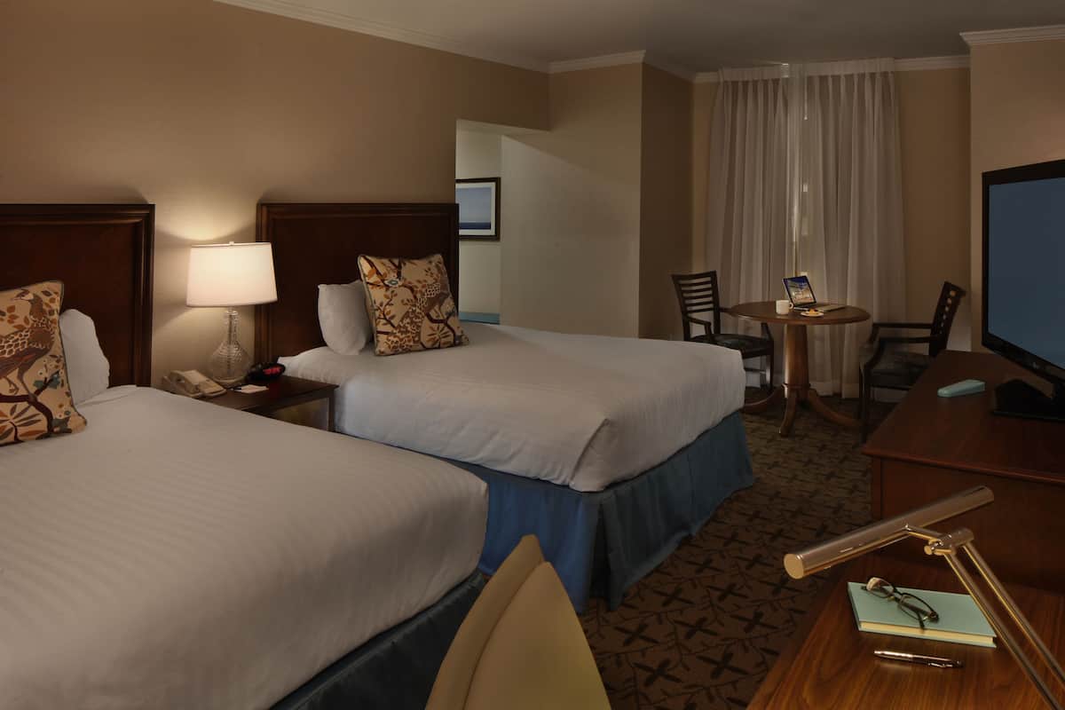 A hotel room with two beds, a desk, a chair, a lamp, a television, and a window with curtains.