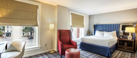 A hotel room with a bed, a chair, a table, and a window with a view of the street.