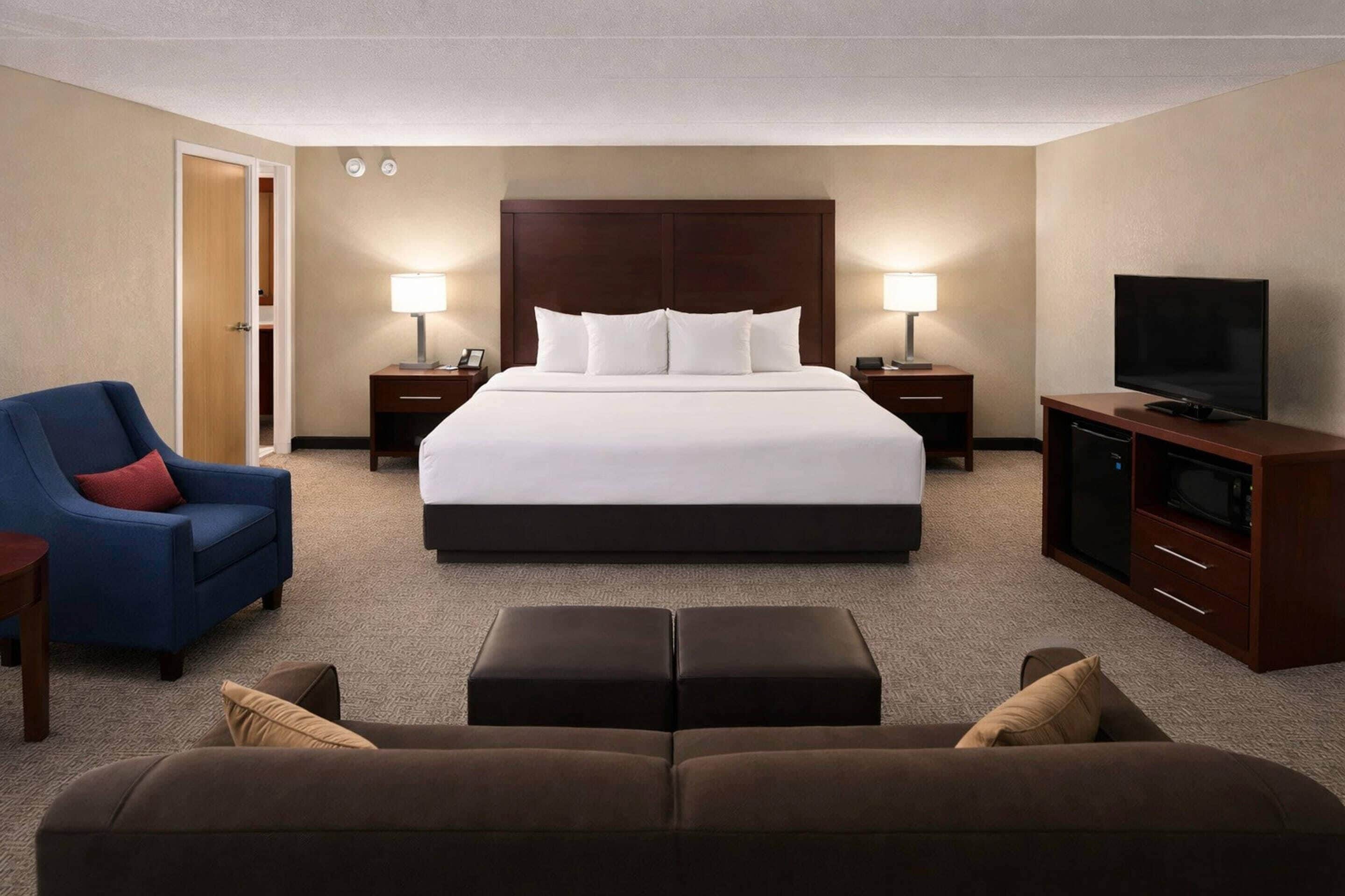 A hotel room with a large bed, two bedside tables with lamps, a television, a blue armchair, and a brown sofa.