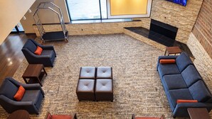 A spacious lounge area with a central ottoman, sofas, and armchairs, a fireplace, and a television mounted on the wall. - Comfort Inn  Farmington Hills - Detroit Northwest (Farmington Hills)